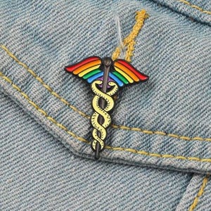 May include: A colorful enamel pin featuring the Caduceus symbol, with rainbow-colored wings and a yellow serpent entwined around a staff. The pin is set against a denim jacket.