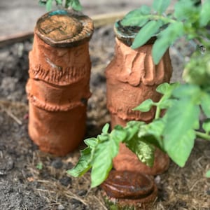 Könnte beinhalten: Zwei terrakottafarbene Keramik-Gartentöpfe mit strukturierten Oberflächen und dunkelbraunen Oberseiten. Die Töpfe sind zylindrisch mit einem geriffelten Design. Grüne Blätter einer nahegelegenen Pflanze verdecken teilweise die Sicht.