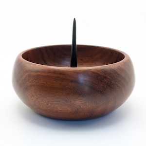 May include: A handcrafted wooden bowl with a dark finish. A tall, slender, black spike rises from the center. The bowl's rich, warm tones and smooth texture suggest a decorative or functional item for the home.