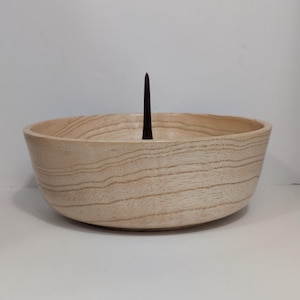 May include: A light-colored wooden bowl with a dark brown, pointed object rising from the center. The bowl has a smooth, rounded shape and visible wood grain. The object appears to be a decorative element or a holder.