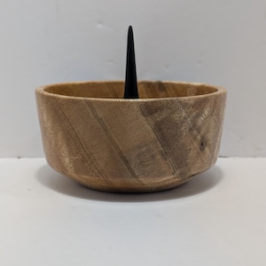 May include: A wooden bowl with a dark, conical object rising from the center. The bowl is light brown with visible wood grain. The dark object is black and tapers to a point. The bowl is round and has a simple, natural design.