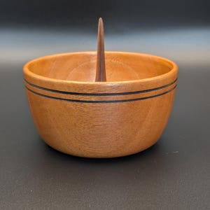 May include: A wooden bowl with a dark brown finish and two black stripes near the top rim. A tall, thin, wooden spike rises from the center of the bowl. The bowl is likely for decorative or functional use.