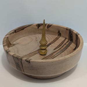 May include: A handcrafted wooden bowl with a light brown finish and dark streaks. A decorative, golden-colored, wooden spire stands in the center of the bowl. The bowl is round and has a smooth, polished surface.