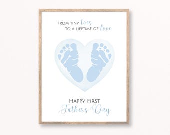 May include: A framed print with a light wood frame. The print features a heart-shaped design with baby footprints in light blue. Text at the top reads "From tiny toes to a lifetime of love." Below the heart, the text reads "Happy First Father's Day."