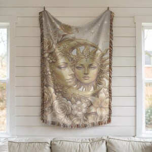 May include: A beige woven throw blanket featuring a celestial design. The artwork depicts two female faces, one within a crescent moon and the other crowned with sun rays, surrounded by flowers. The blanket has a fringed edge.
