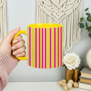 May include: A yellow coffee mug with a matching yellow handle and interior. The mug's exterior features vertical stripes in bright pink and white against a light green background. The mug is held by a person.