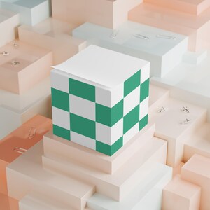 Puede incluir: Un bloc de notas blanco en forma de cubo con un patrón de tablero de ajedrez verde y blanco en los lados. El bloc de notas se encuentra sobre una serie de formas geométricas de colores pastel, con clips esparcidos.
