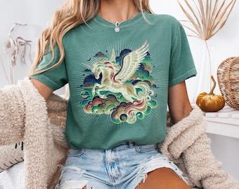 May include: A light green short-sleeve t-shirt featuring a graphic print of a winged horse, or pegasus, surrounded by stylized clouds and radiating light lines. The shirt has a crew neckline and a relaxed fit, worn with light blue denim shorts.