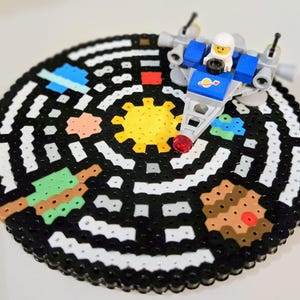 May include: A black and white bead board with a colorful depiction of a solar system. A small blue and gray spaceship with a red light sits on top of the board.