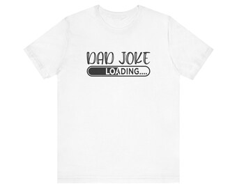 May include: A white short-sleeved t-shirt featuring black text that reads DAD JOKE LOADING with a partial progress bar graphic underneath the text. The design is centered on the chest area of the shirt, which has a crew neckline.