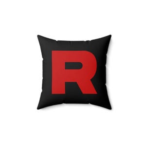 May include: Square black throw pillow with a large red letter "R" in the center. The pillow is a solid black color with a bold red letter, creating a striking contrast. The pillow is a decorative item.