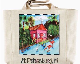 May include: A beige canvas tote bag with a colorful illustration of a pink house with a porch, palm trees, and flamingos on a blue background. The text "St. Petersburg, FL" is printed on the bag.