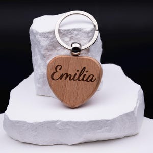 Einzelner Herz Schlüsselanhänger aus Holz mit Gravur des Namens Emilia. Das naturbelassene Material und die klare Schrift verleihen dem Anhänger eine edle Optik. Personalisierbar mit jedem Namen oder Motiv ist er eine schöne Geschenkidee für Geburtstage, Jubiläen, Valentinstag oder als kleine Aufmerksamkeit. Robust und langlebig, zugleich stilvoll und modern als Alltagsaccessoire.