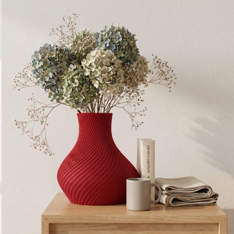 May include: A red textured vase holds a bouquet of dried flowers with blue, green, and beige hues. A small gray mug, a folded beige cloth, and a book are on a wooden surface, creating a still-life arrangement.