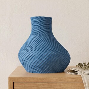 May include: A blue decorative vase with a textured, lattice-like pattern. The vase has a wide, rounded base that tapers to a narrower neck and opening. It sits on a wooden surface, with a folded cloth and sprig of greenery nearby.