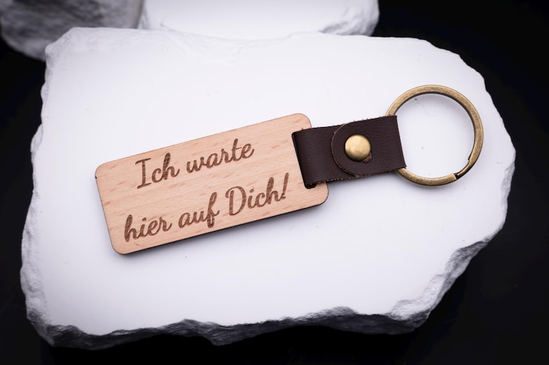 Ein gravierter Schlüsselanhänger aus hellem Holz mit der Aufschrift „Ich warte hier auf Dich!“ liegt auf weißem Stein. Er ist mit einem stabilen Metallring in Antik-Gold und braunem Lederband ausgestattet. Der Anhänger bietet Platz für persönliche Gravuren und individuelle Sprüche, ideal für besondere Geschenke mit emotionalem Wert. Praktisch und stilvoll, ein Begleiter für Schlüsselbund, Auto oder Tasche mit persönlicher Note.