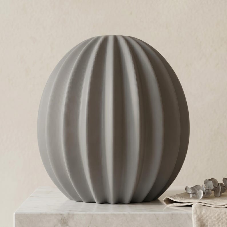 May include: A grey, spherical decorative vase with vertical ribbed detailing. The vase is made of a matte material and sits on a marble surface. The vase has a modern, minimalist design.