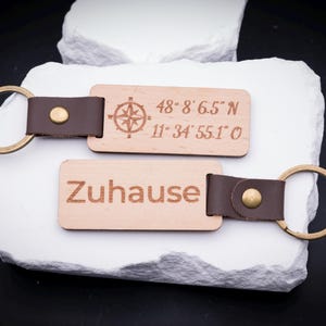 Zwei gravierte Holz Schlüsselanhänger auf weißem Stein, mit den Gravuren „Zuhause“ sowie geografischen Koordinaten inklusive Kompasssymbol. Gefertigt aus hellem Holz mit braunem Lederband und stabilem Metallring in Antik-Optik. Perfekt für personalisierte Gravuren, ob Namen, Sprüche oder Koordinaten. Ein besonderes Geschenk für Familien, Paare oder Freunde, die ihre Schlüssel mit einem stilvollen und individuellen Accessoire aufwerten möchten.