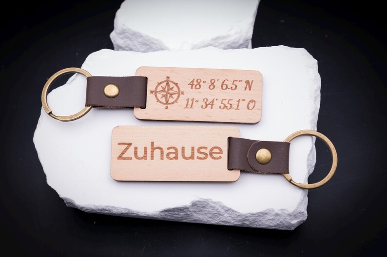 Zwei gravierte Holz Schlüsselanhänger auf weißem Stein, mit den Gravuren „Zuhause“ sowie geografischen Koordinaten inklusive Kompasssymbol. Gefertigt aus hellem Holz mit braunem Lederband und stabilem Metallring in Antik-Optik. Perfekt für personalisierte Gravuren, ob Namen, Sprüche oder Koordinaten. Ein besonderes Geschenk für Familien, Paare oder Freunde, die ihre Schlüssel mit einem stilvollen und individuellen Accessoire aufwerten möchten.