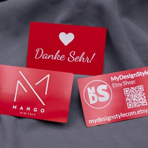 Drei rote Visitenkarten aus Aluminium auf grauem Stoff. Oben eine Dankeskarte mit Herz und Gravur „Danke Sehr!“, unten links das Mango-Digitals-Logo und rechts die MyDesignStyle-Karte mit QR-Code und Etsy-Shop-Adresse. Die Metallkarten haben eine matte Oberfläche, sind langlebig und hochwertig – perfekt als exklusive Businesskarten, Kundendankeskarten oder Werbemittel im modernen Design.