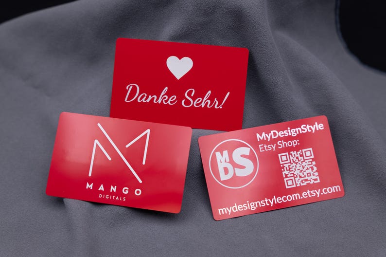 Drei rote Visitenkarten aus Aluminium auf grauem Stoff. Oben eine Dankeskarte mit Herz und Gravur „Danke Sehr!“, unten links das Mango-Digitals-Logo und rechts die MyDesignStyle-Karte mit QR-Code und Etsy-Shop-Adresse. Die Metallkarten haben eine matte Oberfläche, sind langlebig und hochwertig – perfekt als exklusive Businesskarten, Kundendankeskarten oder Werbemittel im modernen Design.