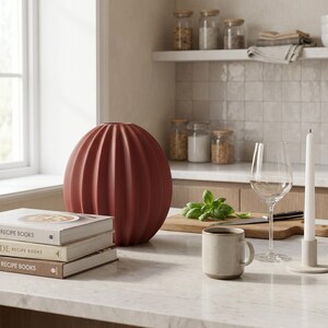 May include: A terracotta-coloured, ribbed ceramic vase sits on a marble countertop alongside recipe books. A wine glass, a mug, and a candle holder with a lit candle are also present. A wooden chopping board with basil and a knife is in the background.