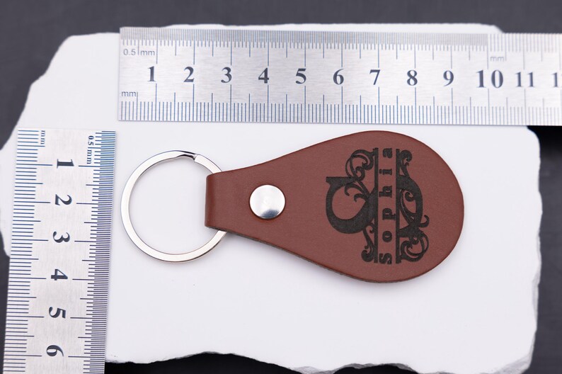Personalisierter Schlüsselanhänger aus braunem Kunstleder mit Gravur und Maßangaben zur Orientierung. Die Lineale zeigen eine Länge von etwa 9 cm und eine Breite von rund 4,5 cm – kompakt, aber dennoch gut sichtbar. Der Anhänger vereint elegantes Design, individuelle Gravur und Funktionalität und eignet sich perfekt für Autoschlüssel, Haustürschlüssel oder als personalisiertes Geschenk.