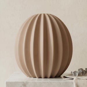 May include: A beige, spherical decorative vase with vertical, ribbed detailing. The vase has a rounded shape and a neutral colour, suitable for various interior design styles. It is placed on a marble surface.