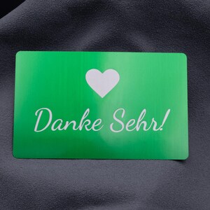 Grüne Metall-Visitenkarte mit lasergraviertem Herzsymbol und Schriftzug „Danke Sehr!“. Die Karte wirkt modern und stilvoll, ideal als hochwertige Beigabe zu Bestellungen, als Kundendankeschön oder zur Markenbindung. Die Aluminiumoberfläche ist langlebig, stabil und vermittelt Exklusivität, wodurch sich diese Karte von herkömmlichen Papierkarten deutlich abhebt.