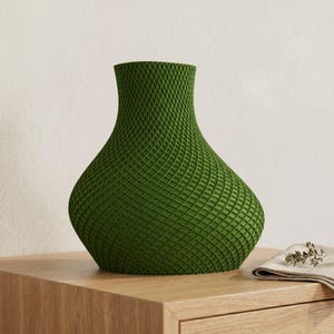 May include: A green decorative vase with a lattice pattern. The vase has a wide base that tapers to a narrow neck. It is sitting on a wooden surface. A small linen cloth with sprigs of herbs is next to the vase.