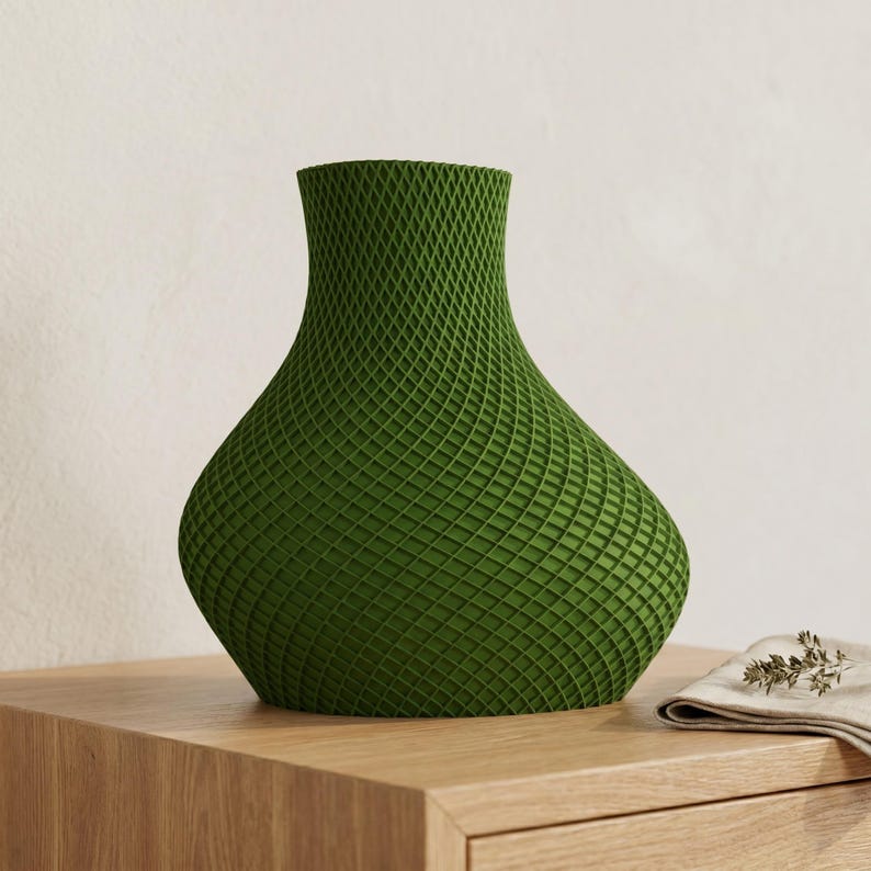 May include: A green decorative vase with a lattice pattern. The vase has a wide base that tapers to a narrow neck. It is sitting on a wooden surface. A small linen cloth with sprigs of herbs is next to the vase.