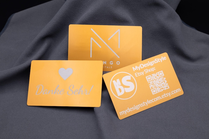 Drei Metall-Visitenkarten in Hellgold mit Gravur: eine Dankeskarte mit Herzsymbol und „Danke Sehr!“, eine Mango-Digitals-Karte mit Logo sowie eine MyDesignStyle-Karte mit QR-Code und Etsy-Shop-Adresse. Die robusten Aluminiumkarten mit matter Oberfläche und abgerundeten Ecken vereinen modernes Design mit Funktionalität und eignen sich perfekt als hochwertige Visitenkarten, Kundenkarten oder elegante Dankeskarten.
