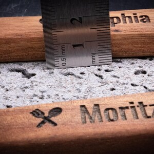 Detailaufnahme des gravierten Kochlöffelsets aus Akazienholz mit Edelstahl-Lineal zur Messung der Dicke. Das Maß zeigt etwa 1,5 cm Stärke des robusten Holzes. Sichtbar sind die Gravuren „Sophia wendet mit Liebe!“ und „Moritz rührt mit Gefühl!!!“ mit kleinen Symbolen. Das hochwertige Akazienholz mit feiner Maserung und präziser Lasergravur steht für langlebige Qualität – ideal als personalisiertes Küchenutensil oder Geschenk.
