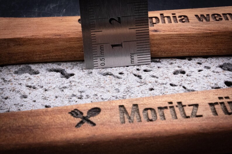 Detailaufnahme des gravierten Kochlöffelsets aus Akazienholz mit Edelstahl-Lineal zur Messung der Dicke. Das Maß zeigt etwa 1,5 cm Stärke des robusten Holzes. Sichtbar sind die Gravuren „Sophia wendet mit Liebe!“ und „Moritz rührt mit Gefühl!!!“ mit kleinen Symbolen. Das hochwertige Akazienholz mit feiner Maserung und präziser Lasergravur steht für langlebige Qualität – ideal als personalisiertes Küchenutensil oder Geschenk.