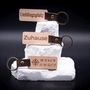 Drei gravierte Holz Schlüsselanhänger sind dekorativ aufgestapelt, versehen mit den Gravuren „Lieblingsplatz“, „Zuhause“ sowie geografischen Koordinaten mit Kompasssymbol. Die Anhänger bestehen aus hellem Holz, kombiniert mit braunem Lederband und massivem Metallring in Antik-Optik. Ein stilvolles Accessoire für Schlüsselbund oder Auto, perfekt als personalisiertes Geschenk für Paare, Familie oder Freunde mit individuellem Wunschtext oder Koordinaten.