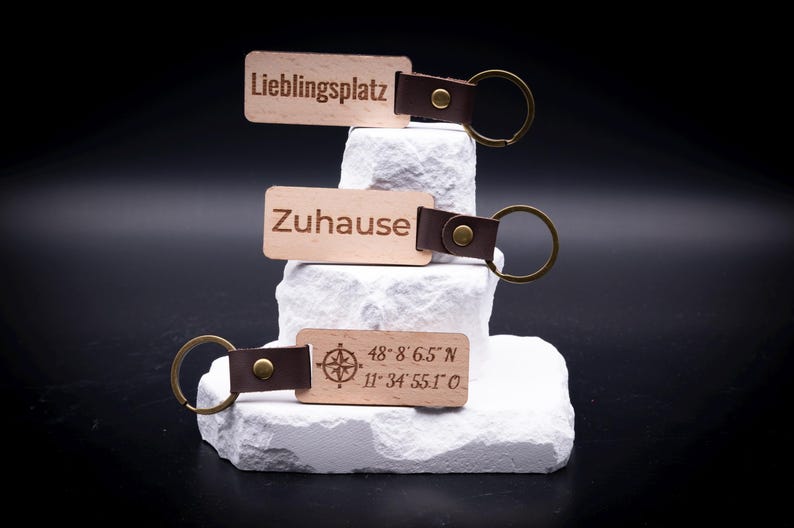 Drei gravierte Holz Schlüsselanhänger sind dekorativ aufgestapelt, versehen mit den Gravuren „Lieblingsplatz“, „Zuhause“ sowie geografischen Koordinaten mit Kompasssymbol. Die Anhänger bestehen aus hellem Holz, kombiniert mit braunem Lederband und massivem Metallring in Antik-Optik. Ein stilvolles Accessoire für Schlüsselbund oder Auto, perfekt als personalisiertes Geschenk für Paare, Familie oder Freunde mit individuellem Wunschtext oder Koordinaten.