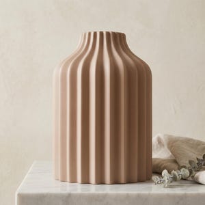 Może przedstawiać: Beżowy ceramiczny wazon z żebrowanym wzorem. Wazon ma cylindryczny korpus i lekko rozszerzającą się szyję. Wazon stoi na białej marmurowej powierzchni. Beżowa tkanina i trochę zieleni są w tle.