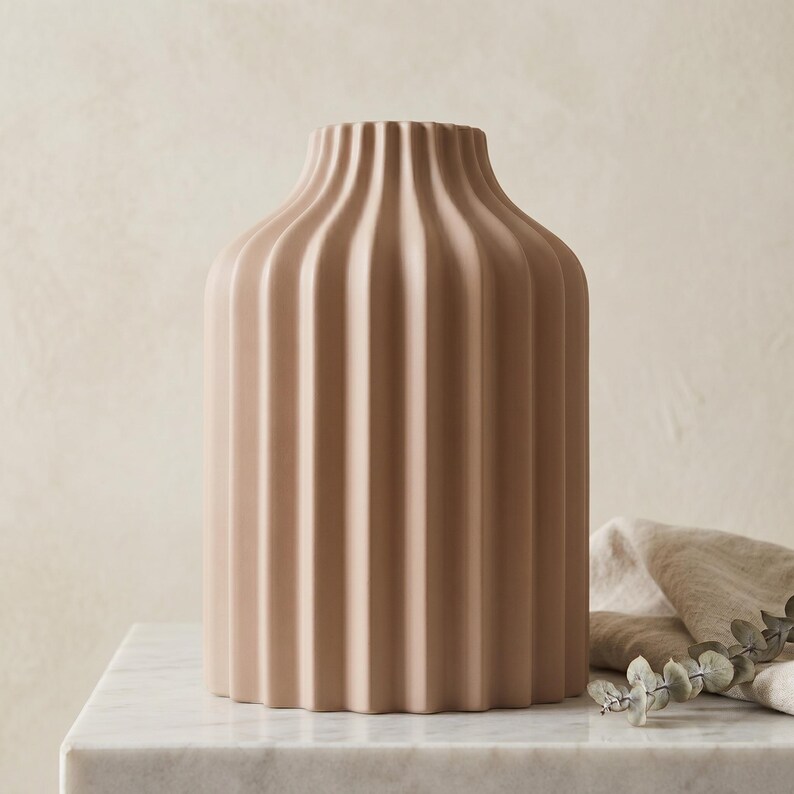 Może przedstawiać: Beżowy ceramiczny wazon z żebrowanym wzorem. Wazon ma cylindryczny korpus i lekko rozszerzającą się szyję. Wazon stoi na białej marmurowej powierzchni. Beżowa tkanina i trochę zieleni są w tle.