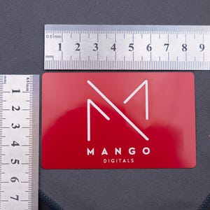 Rote Metallkarte mit Mango-Digitals-Logo, dargestellt mit Lineal zur Größenangabe. Das Format entspricht einer klassischen Kreditkarte (8,6 × 5,4 cm). Langlebig, stabil und edel – ideal als exklusive Businesskarte oder kreative Werbekarte.
