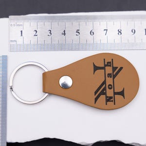 Personalisierter Schlüsselanhänger aus hellbraunem veganem Kunstleder mit Gravur „Noah“ im Monogramm-Stil. Daneben liegt ein Maßband aus Edelstahl, das die Größe des Anhängers verdeutlicht. Die Kombination aus Naturton und schwarzer Gravur macht das Accessoire zu einem modernen, personalisierten Geschenk für Männer, Frauen oder Paare.