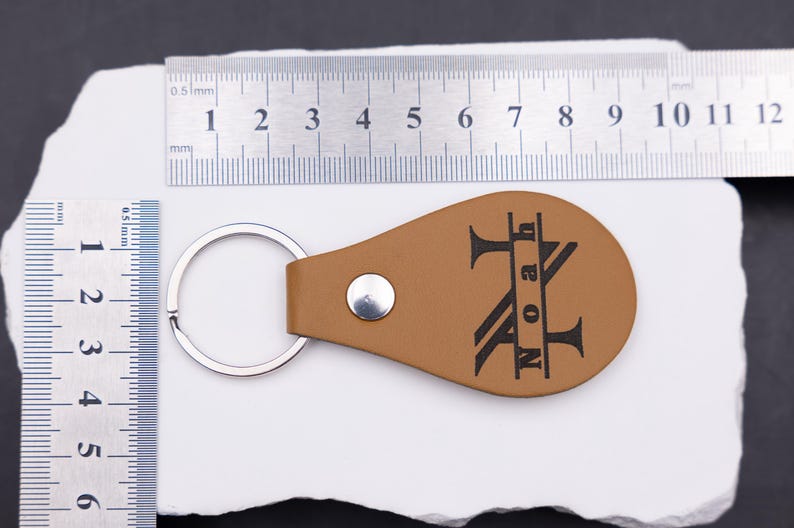 Personalisierter Schlüsselanhänger aus hellbraunem veganem Kunstleder mit Gravur „Noah“ im Monogramm-Stil. Daneben liegt ein Maßband aus Edelstahl, das die Größe des Anhängers verdeutlicht. Die Kombination aus Naturton und schwarzer Gravur macht das Accessoire zu einem modernen, personalisierten Geschenk für Männer, Frauen oder Paare.