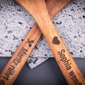 Eng gefasste Aufnahme des 2er-Kochlöffelsets mit Gravuren „Sophia wendet mit Liebe!“ und „Moritz rührt mit Gefühl!!!“. Die dunkle Gravur wirkt präzise und kontrastreich auf dem hellbraunen Akazienholz. Perfekt als personalisiertes Geschenkset für Küche, Grill oder Backfreunde.
