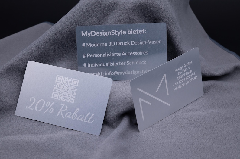 Drei exklusive Silber-Visitenkarten aus Aluminium, präsentiert auf grauem Stoff. Links eine Karte mit Gravur „20% Rabatt“ und QR-Code, in der Mitte eine Informationskarte mit Details zu personalisierten Produkten, rechts eine klassische Businesskarte mit Firmenlogo und Kontaktinformationen. Die Metallkarten wirken edel und robust, ideal für Business, Werbung und Kundengeschenke im modernen Design.