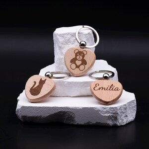 Drei gravierte Schlüsselanhänger in Herzform aus hellem Holz, verziert mit individuellen Motiven und Gravuren. Ein Anhänger zeigt einen Teddybär, ein weiterer eine Katze und der dritte den Namen Emilia in eleganter Schrift. Die Anhänger sind personalisierbar mit Namen oder Symbolen und eignen sich als liebevolles Geschenk für Kinder, Freunde, Partner oder Familie. Nachhaltiges Accessoire mit modernem Design.
