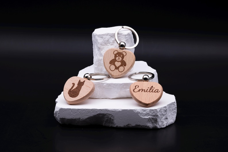 Drei gravierte Schlüsselanhänger in Herzform aus hellem Holz, verziert mit individuellen Motiven und Gravuren. Ein Anhänger zeigt einen Teddybär, ein weiterer eine Katze und der dritte den Namen Emilia in eleganter Schrift. Die Anhänger sind personalisierbar mit Namen oder Symbolen und eignen sich als liebevolles Geschenk für Kinder, Freunde, Partner oder Familie. Nachhaltiges Accessoire mit modernem Design.