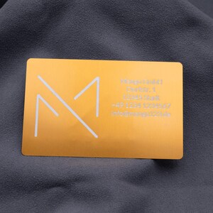 isitenkarte aus Aluminium in Hellgold mit eingraviertem Mango-Logo und detaillierten Kontaktdaten auf der rechten Seite: Adresse, Telefonnummer und E-Mail. Die matte, gebürstete Oberfläche mit abgerundeten Ecken vermittelt einen modernen und professionellen Eindruck. Diese hochwertige Metallkarte eignet sich hervorragend für Geschäftskunden, Networking-Events und repräsentatives Firmenbranding.