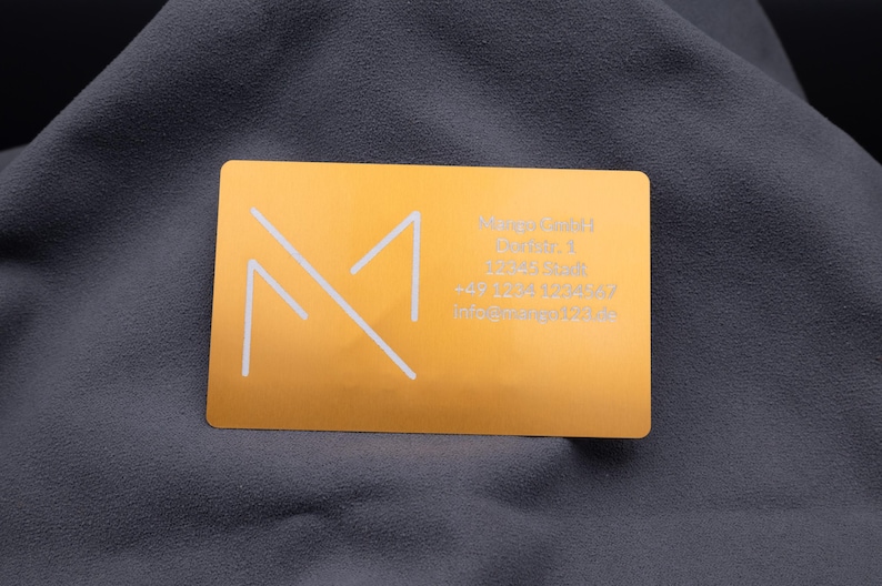 isitenkarte aus Aluminium in Hellgold mit eingraviertem Mango-Logo und detaillierten Kontaktdaten auf der rechten Seite: Adresse, Telefonnummer und E-Mail. Die matte, gebürstete Oberfläche mit abgerundeten Ecken vermittelt einen modernen und professionellen Eindruck. Diese hochwertige Metallkarte eignet sich hervorragend für Geschäftskunden, Networking-Events und repräsentatives Firmenbranding.