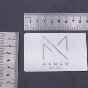 Silberne Visitenkarte aus Aluminium mit graviertem Mango-Logo, dargestellt neben Linealen zur Größenveranschaulichung. Die Maße der Metallkarte entsprechen klassischen Kreditkarten, wodurch sie praktisch und elegant zugleich ist. Ideal als langlebige, exklusive Visitenkarte für Unternehmen, die Wert auf Design, Qualität und auffällige Präsentation legen. Perfekt für Business, Branding und kreative Werbung.