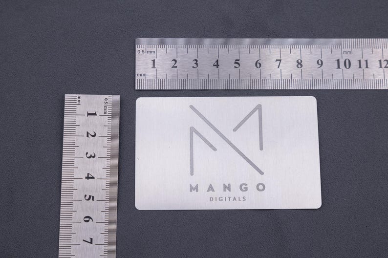 Silberne Visitenkarte aus Aluminium mit graviertem Mango-Logo, dargestellt neben Linealen zur Größenveranschaulichung. Die Maße der Metallkarte entsprechen klassischen Kreditkarten, wodurch sie praktisch und elegant zugleich ist. Ideal als langlebige, exklusive Visitenkarte für Unternehmen, die Wert auf Design, Qualität und auffällige Präsentation legen. Perfekt für Business, Branding und kreative Werbung.