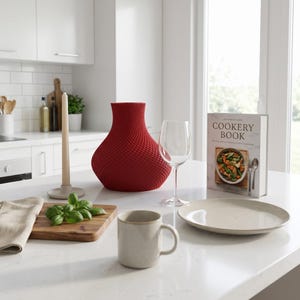 May include: A red textured vase, wine glass, and cookbook on a white countertop. A candle, mug, plate, and cutting board with basil complete the kitchen scene. The cookbook is titled "Cookery Book".